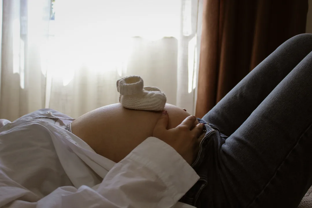 بطن حامل استلقت فوق سرير وفوقه حذاء رضيع صغير